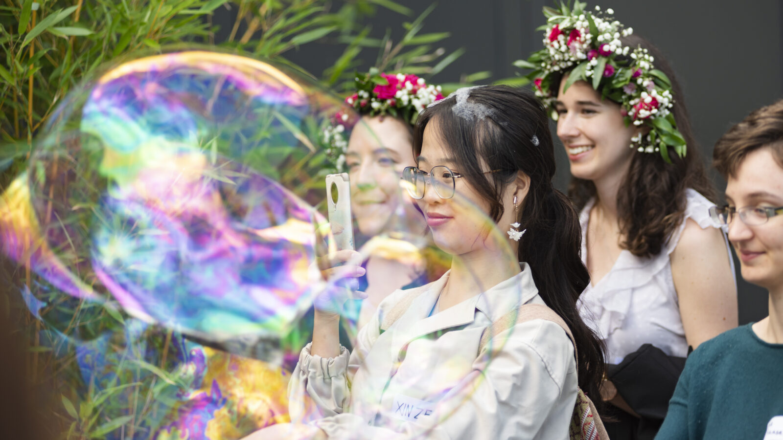Junge Frau macht ein Foto von einer großen Seifenblase. Hinter ihr stehen zwei Frauen mit Blumenkränzen auf dem Kopf und eine Person mit Brille steht rechts von ihr.