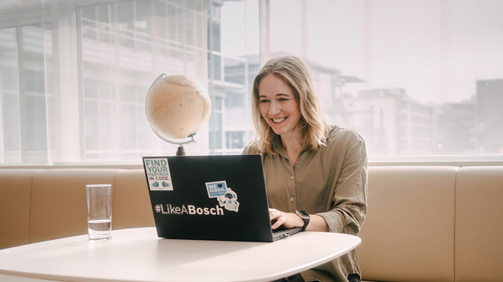 Junge Frau sitzt vor Laptop und schaut auf dessen Bildschirm. Auf dem Laptop sind diverse Sticker zu finden von Bosch. Im Hintergrund ist ein Globus zu sehen und außerhalb des Raumes ein Firmengebäude zu erkennen.