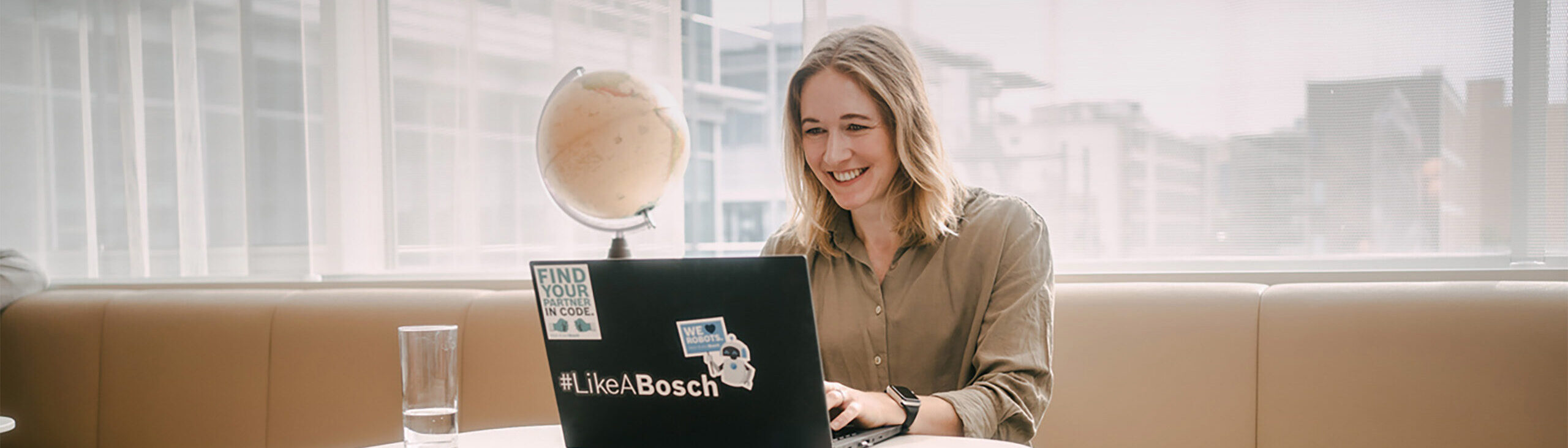 Junge Frau sitzt vor Laptop und schaut auf dessen Bildschirm. Auf dem Laptop sind diverse Sticker zu finden von Bosch. Im Hintergrund ist ein Globus zu sehen und außerhalb des Raumes ein Firmengebäude zu erkennen.