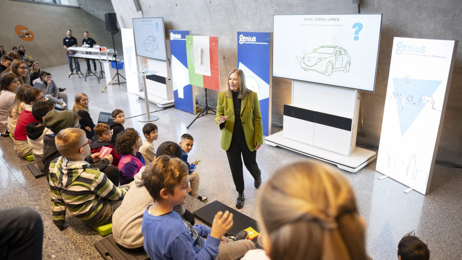 Sabine Kohleisen, die Referentin der Genius Kinderuni, steht auf der Bühne und redet mit den zuschauenden Kindern.